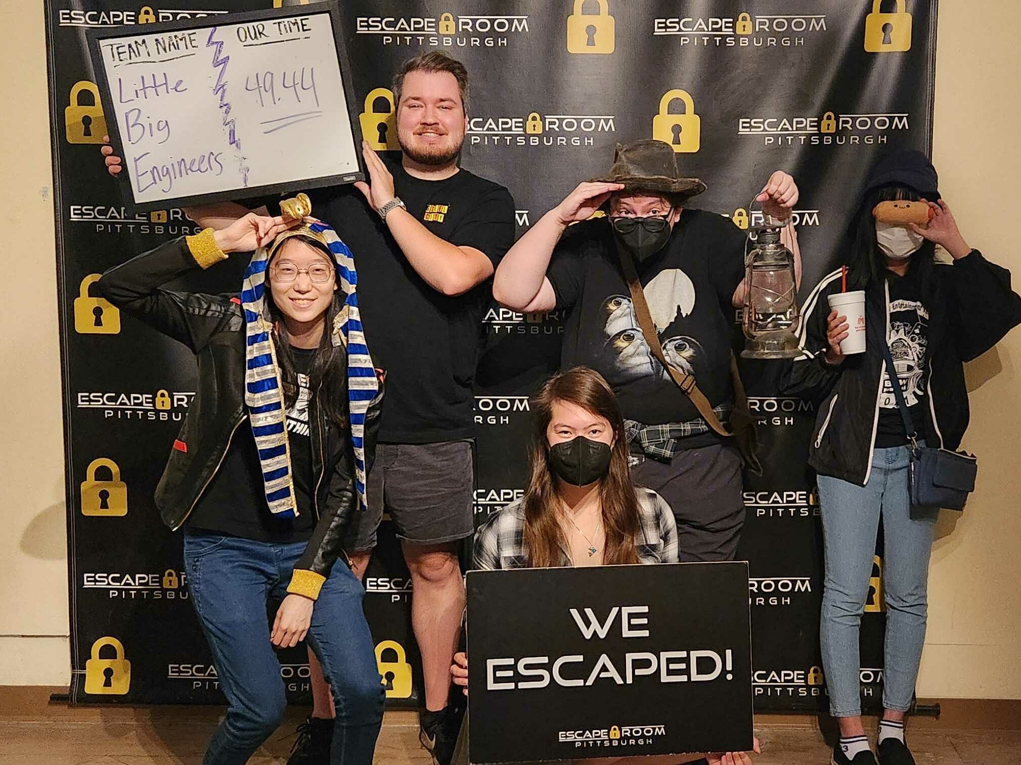 Our team poses in front of the Escape Room Pittsburgh backdrop, wearing items themed to the Tomb Explorer room. Nolan holds a board showing our time of 49:44.